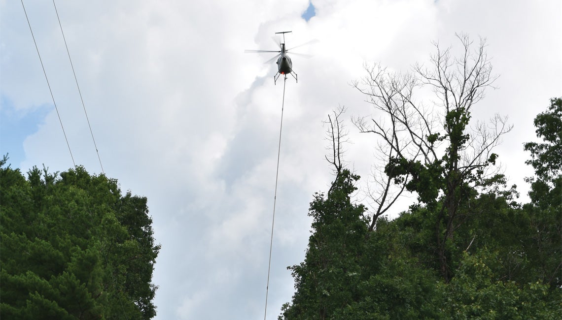 A helicopter flies along a right-of-way in rural Washington County, dangling tree-trimming equipment to remove vegetation near the power lines.