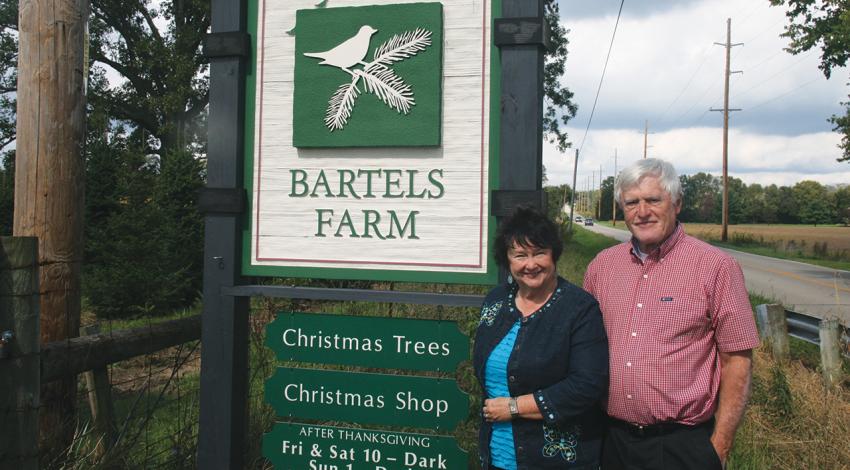 Judy and Steve Bartels smile next to the sign for their farm.