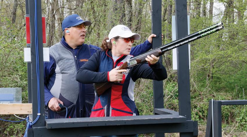 A man stands behind a woman pointing a gun at the clay in the air.