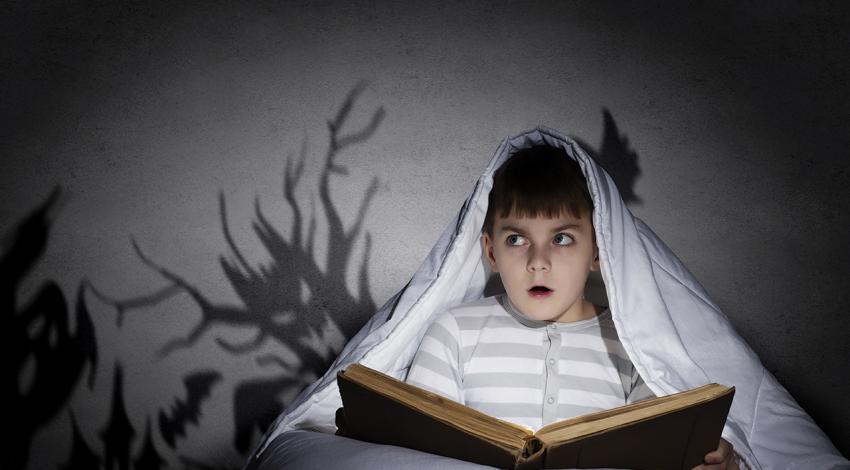 A boy reads under a blanket while scary shadows surround him.