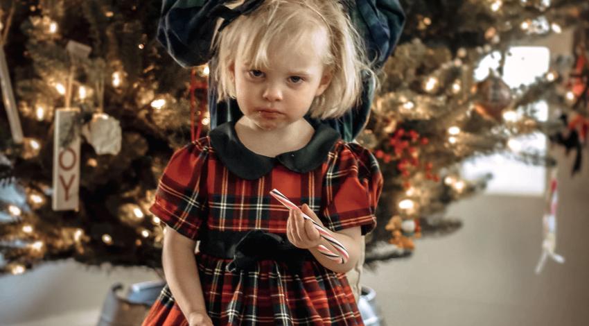Little girl in red dress, holding candy cane, frowns as bigger boy behind her messes up her hair.