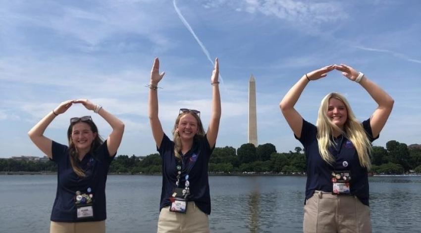 Three teen girls form letters O, H and O with their hands, with obelisk of Washington Monument in the distance, making the I to spell OHIO