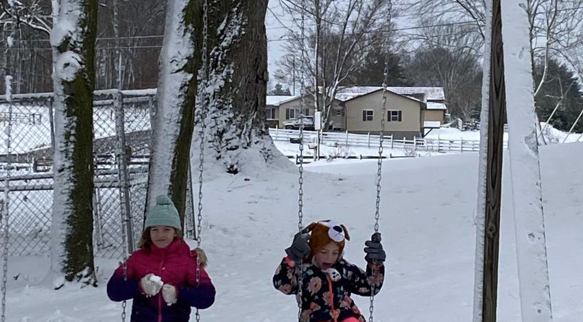 two girls in snow clothes on swings with dog in front