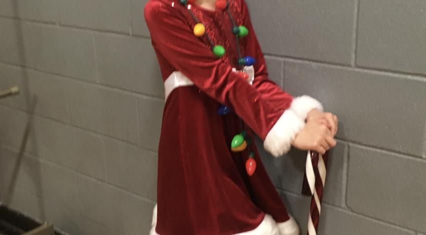 Girl in white-trimmed red dress, wearing Santa hat, leans against cane-sized candy cane