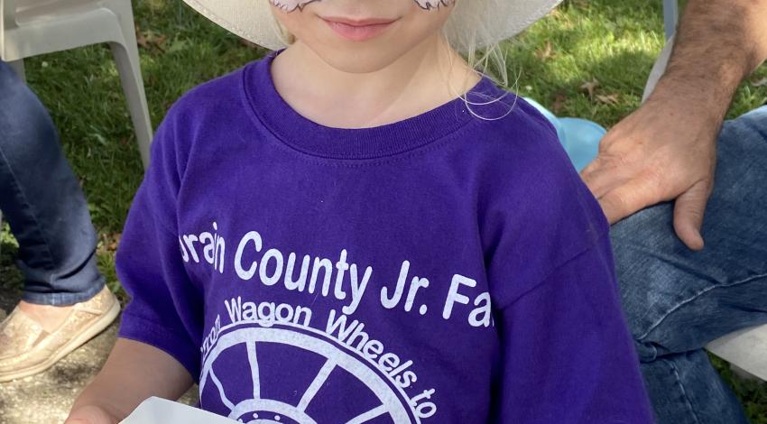 Little girl in cowboy hat, with face painted like a fox, holds corn dog.