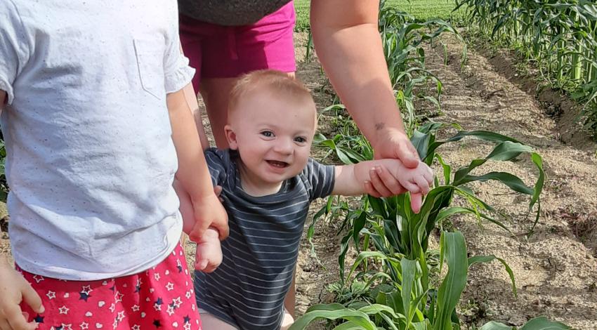 little girl and baby stand in front of mother, next to row of green plants
