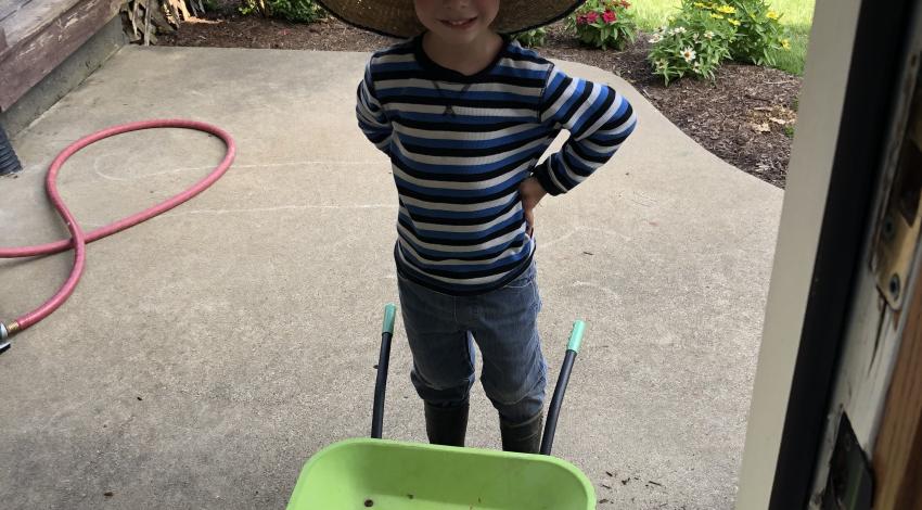 little boy in straw hat stands behind wheelbarrow full of tomatoes and squash