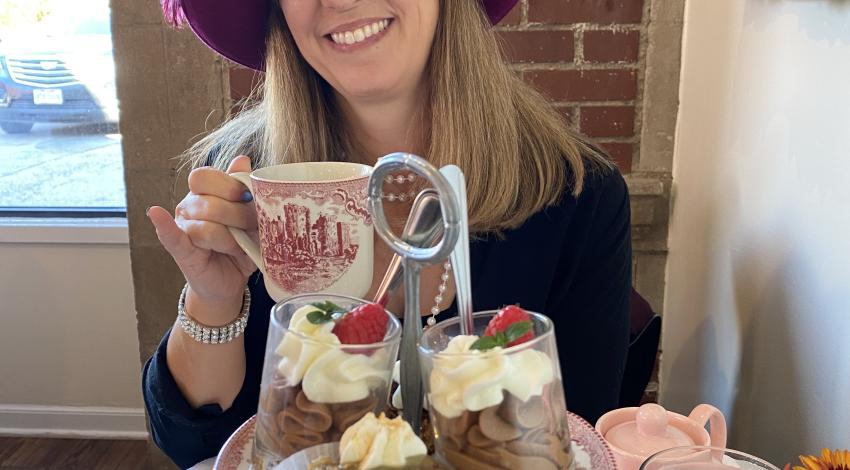 woman in magenta hat with feather holds china teacup behind two-tiered dessert plate