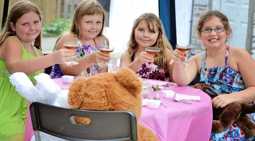 four girls hold up cups like for a toast, across the table from large stuffed rabbit and teddy bear