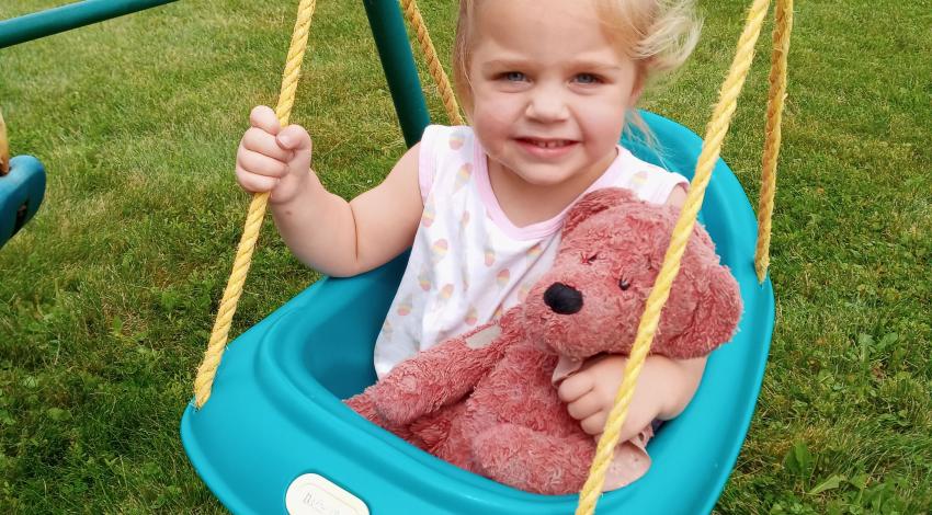 Toddler girl in seat on a swing set, holding a teddy bear