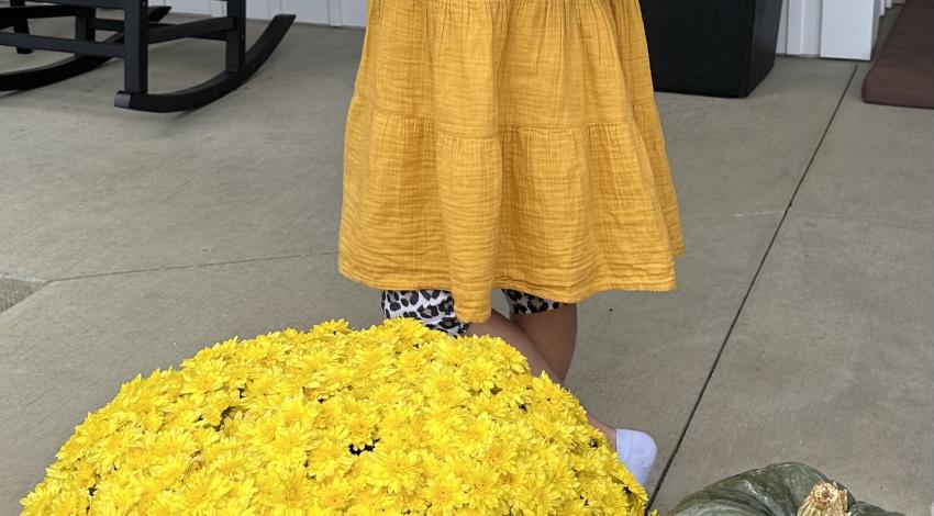 girl in dress behind yellow mum and pumpkin stack