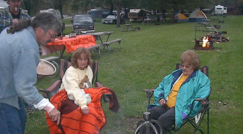 A man stands and two women it near a campfire