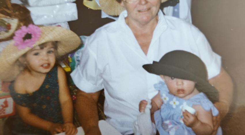 woman sits at table set for tea with little girl and holding baby on her lap, while all three wear straw hats