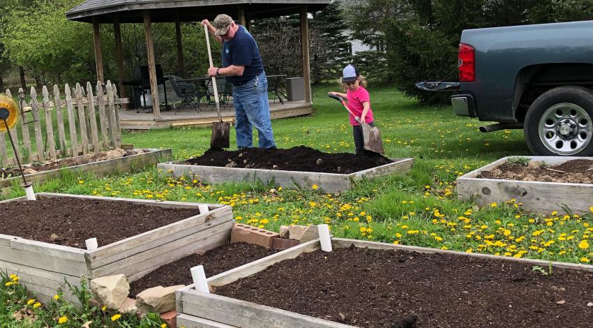 A man and small girl, both wearing hats, shovel soil in wood-framed area