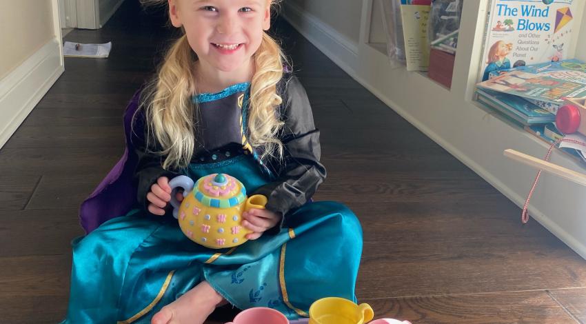 smiling little girl in princess costume and tiara sits cross-legged on the floor behind a tea set, holding a teapot