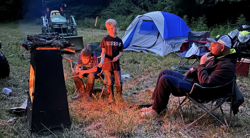 A man and two boys near a fire with a tent in the background.