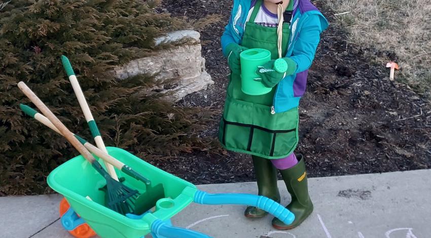 little girl in hat and boots holds watering can beside wheelbarrow