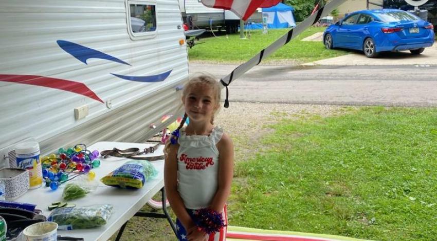 little girl stands under an American flag next to a camper