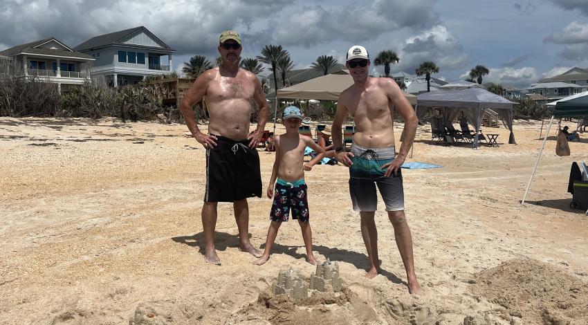 two men and a boy stand behind large sandcastle