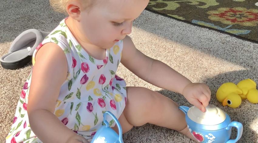 baby girl sits on floor, holding plastic teacup, in sunshine
