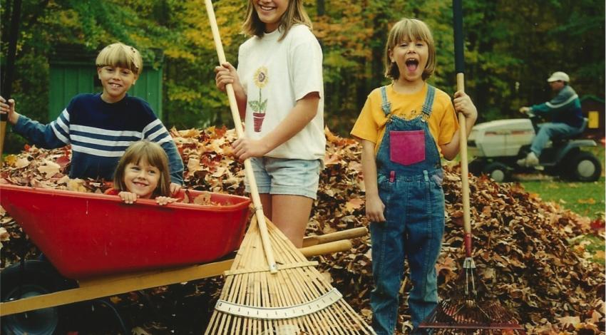 three kids rake leaves with one in wheelbarrow