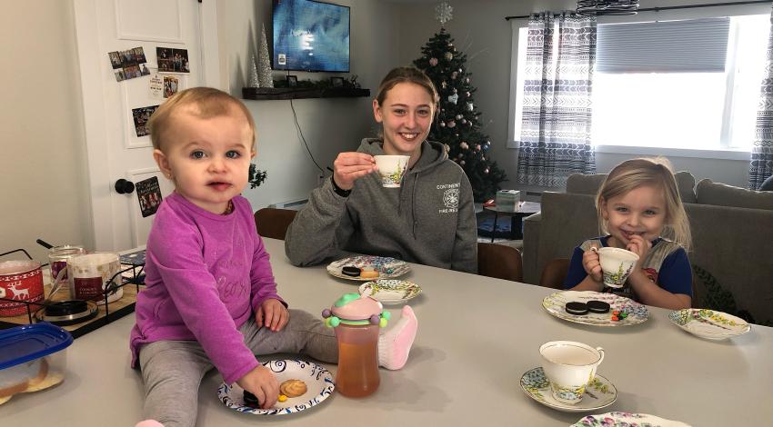 baby sits on table with plate and sippy cup, while little girl and older girl sit behind table holding teacups