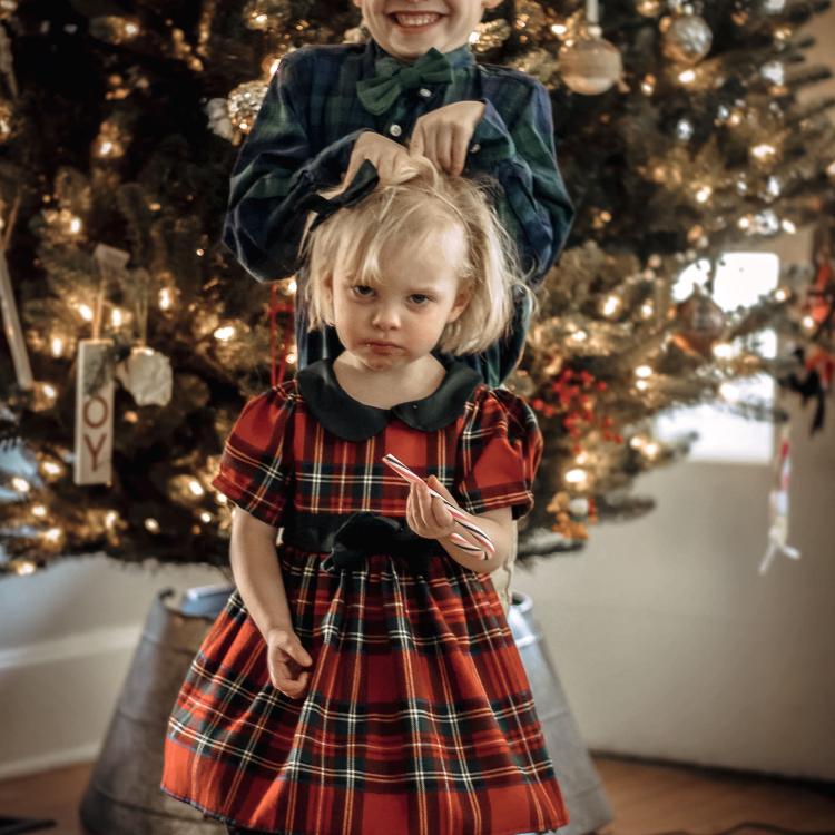 Little girl in red dress, holding candy cane, frowns as bigger boy behind her messes up her hair.