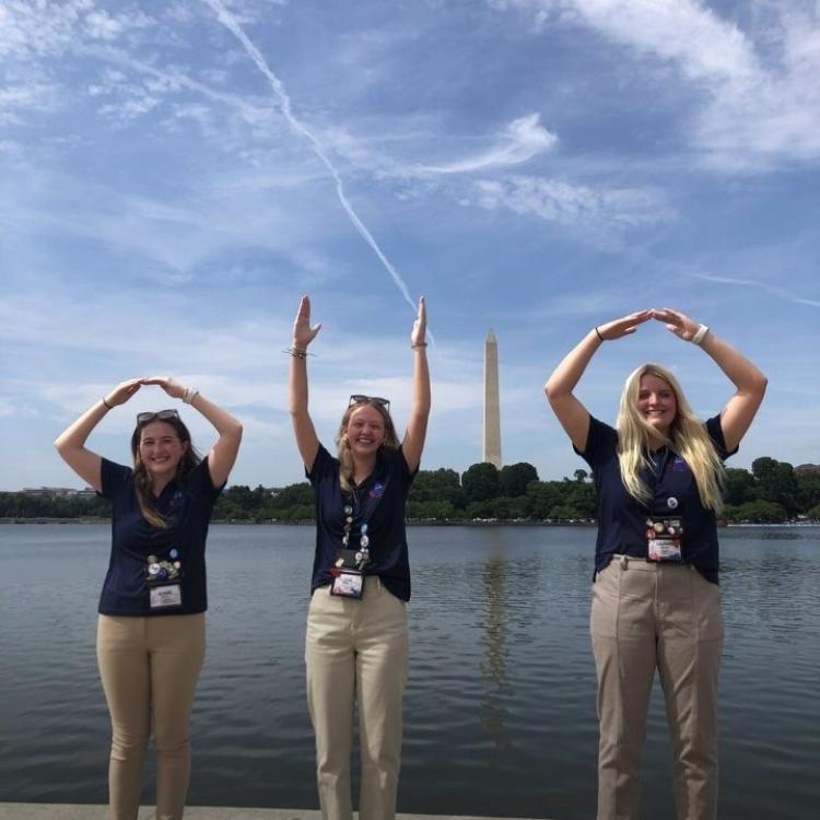 Three teen girls form letters O, H and O with their hands, with obelisk of Washington Monument in the distance, making the I to spell OHIO