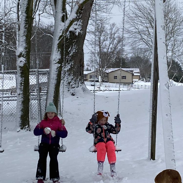 two girls in snow clothes on swings with dog in front