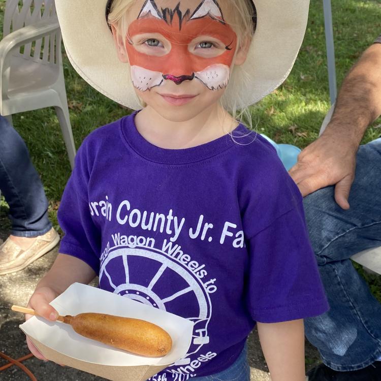 Little girl in cowboy hat, with face painted like a fox, holds corn dog.