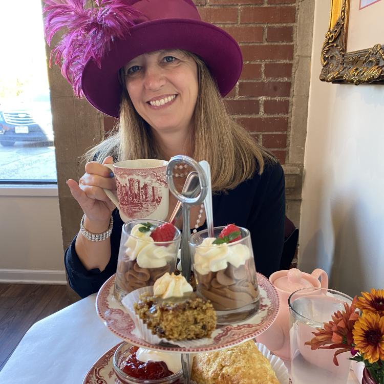 woman in magenta hat with feather holds china teacup behind two-tiered dessert plate