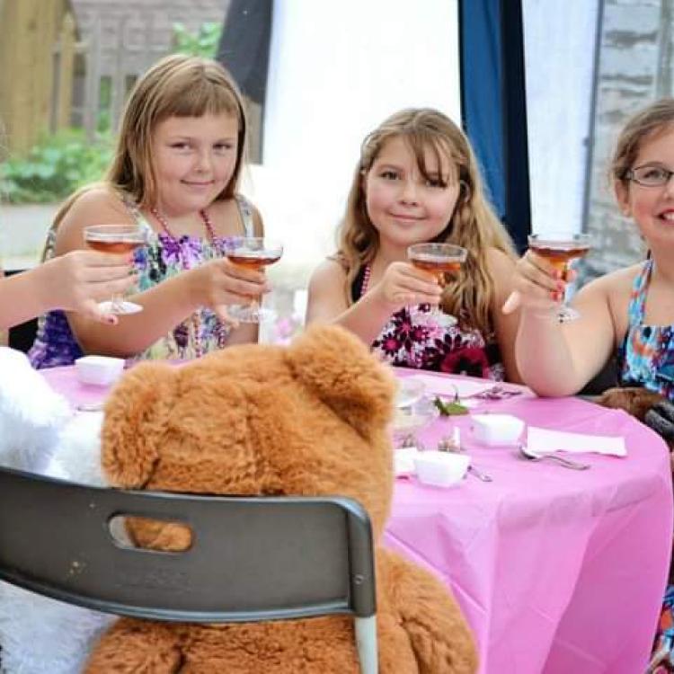 four girls hold up cups like for a toast, across the table from large stuffed rabbit and teddy bear