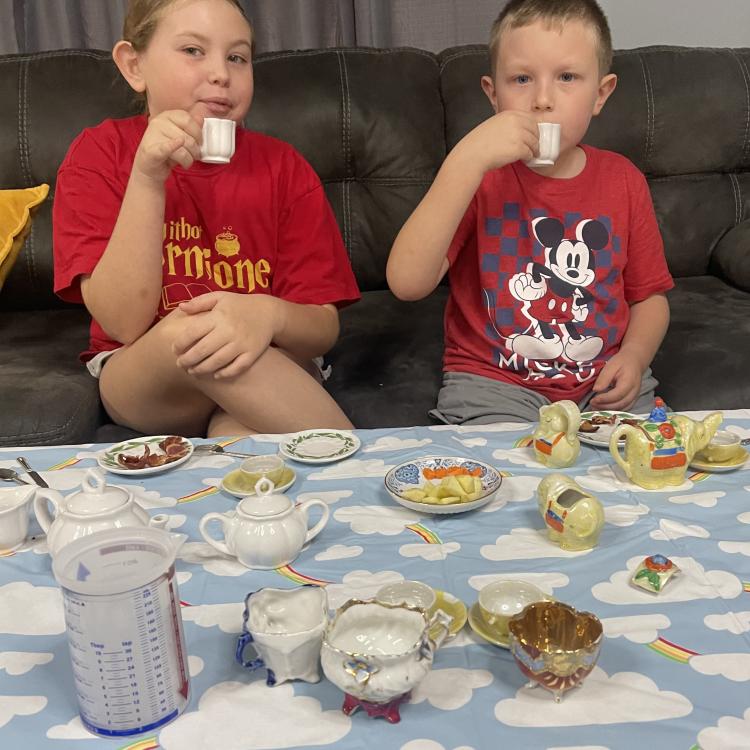 boy and girl sip tea behind table set with china teacups and teapot