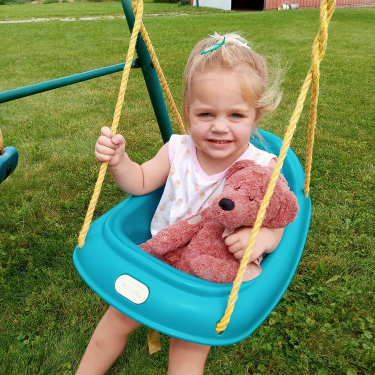 Toddler girl in seat on a swing set, holding a teddy bear