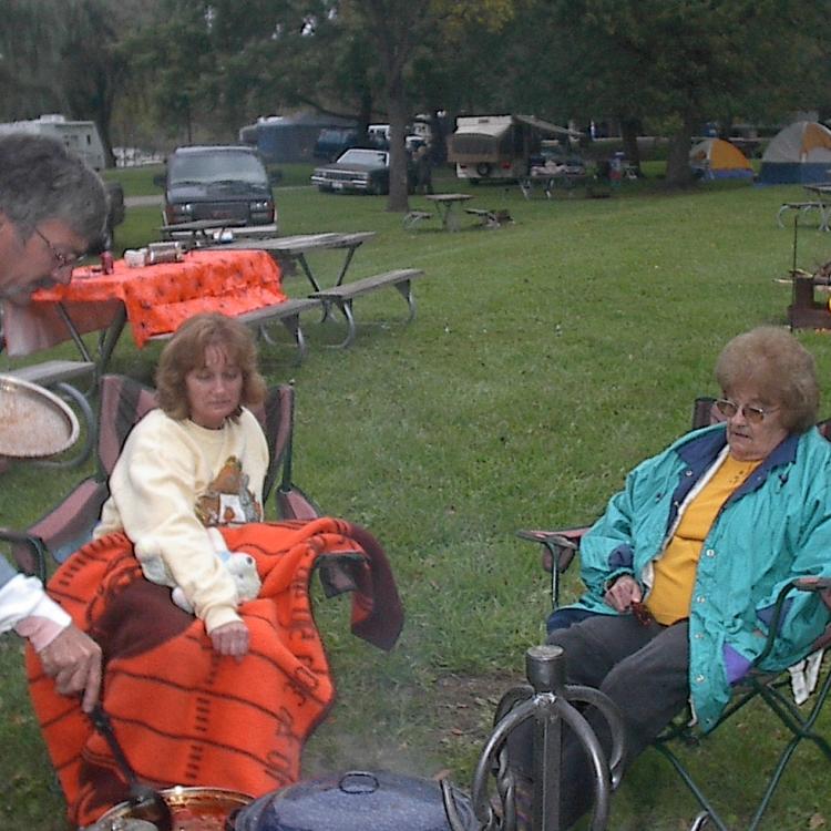 A man stands and two women it near a campfire