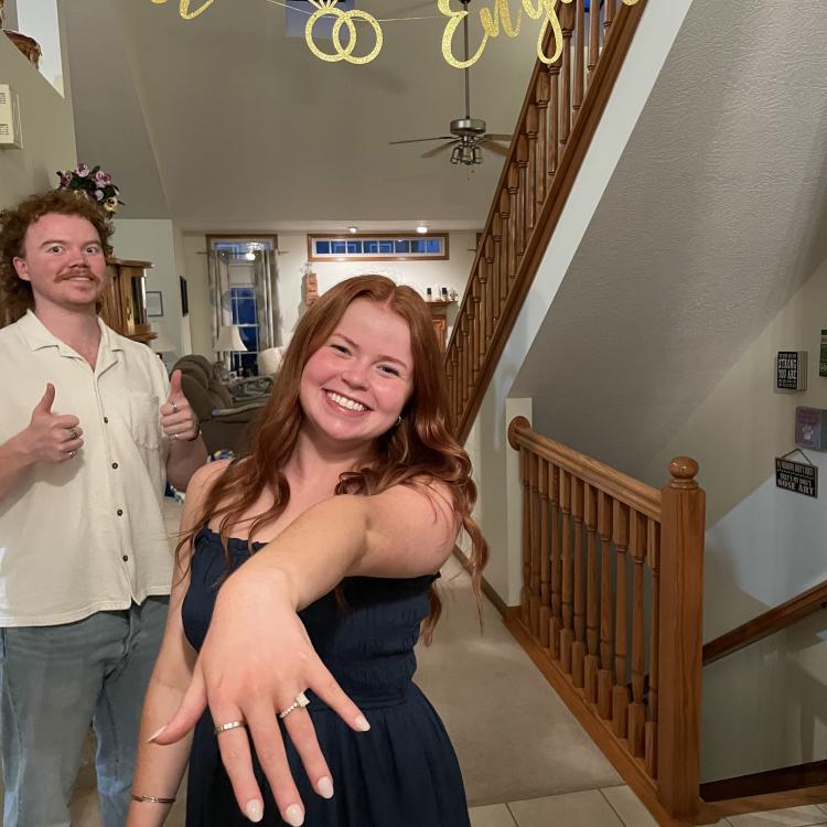 woman shows hand with engagement ring, man in background