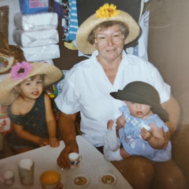 woman sits at table set for tea with little girl and holding baby on her lap, while all three wear straw hats