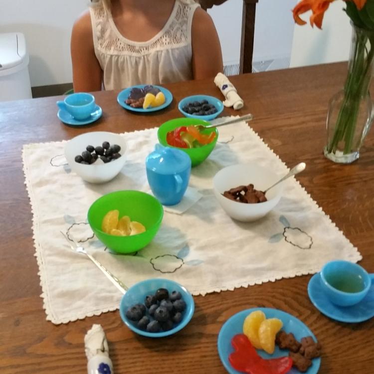 girl sitting behind table set with blue tea set, including cups of tea and fruit on plates