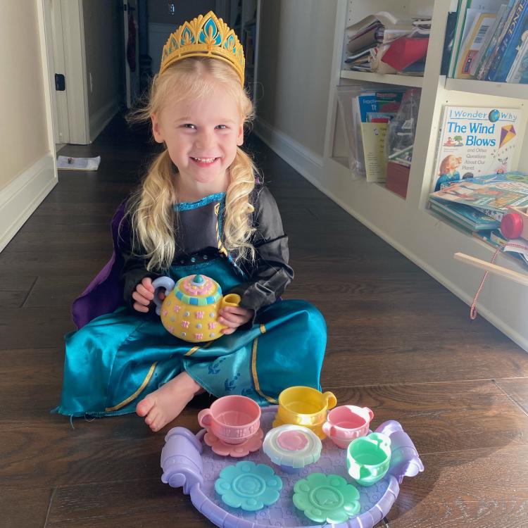 smiling little girl in princess costume and tiara sits cross-legged on the floor behind a tea set, holding a teapot