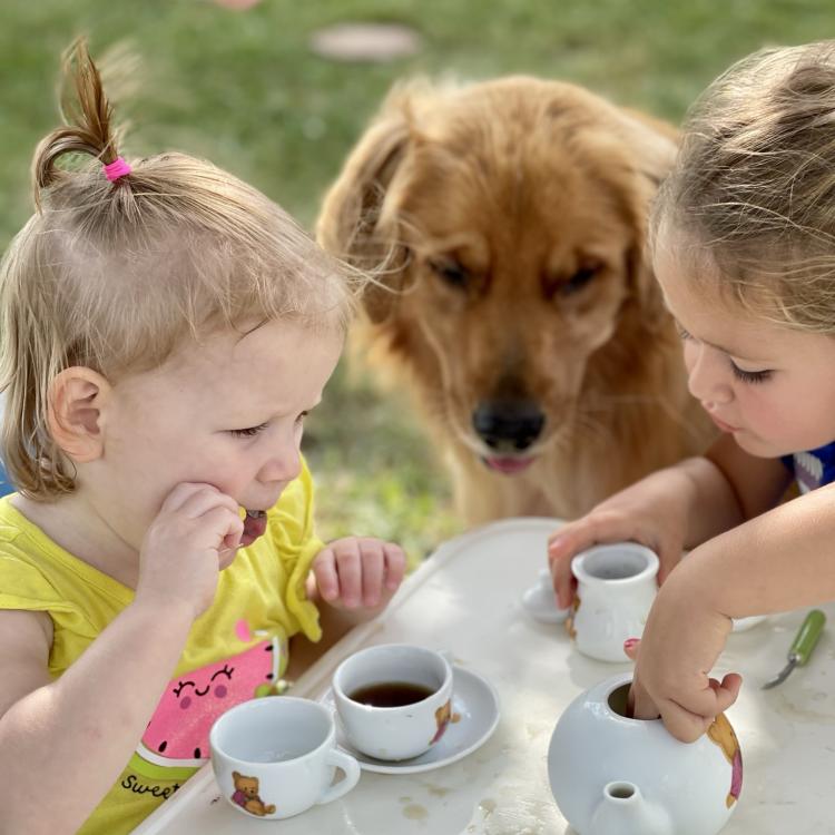two girls sit at outdoor table with tea set while golden retriever sits nearby, watching them