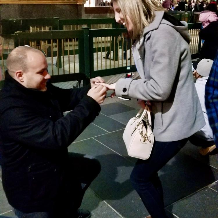 man kneeling in front of woman, putting ring on her finger