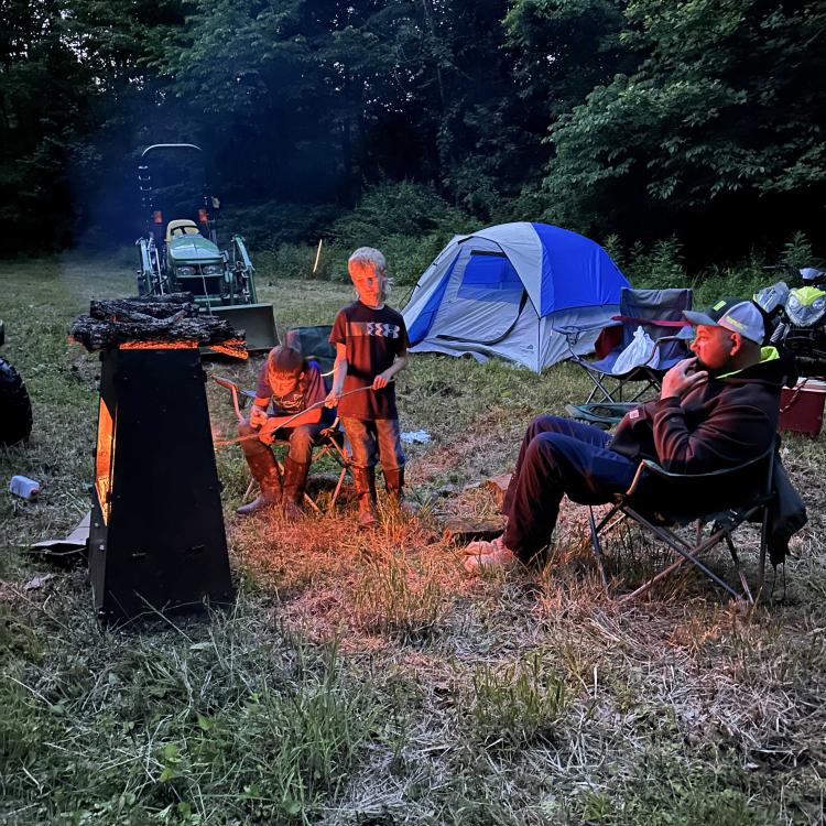 A man and two boys near a fire with a tent in the background.