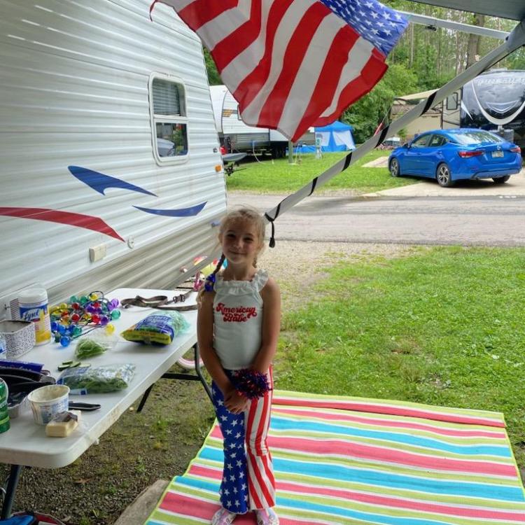 little girl stands under an American flag next to a camper