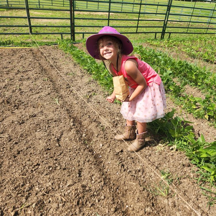 little girl in pink skirt, purple sun hat, and cowgirl boots smiles as she plants seeds