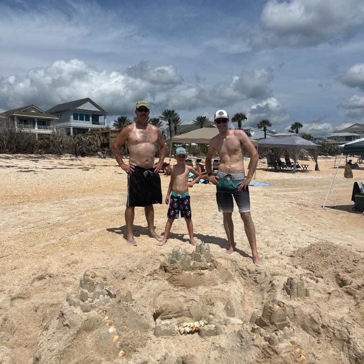 two men and a boy stand behind large sandcastle