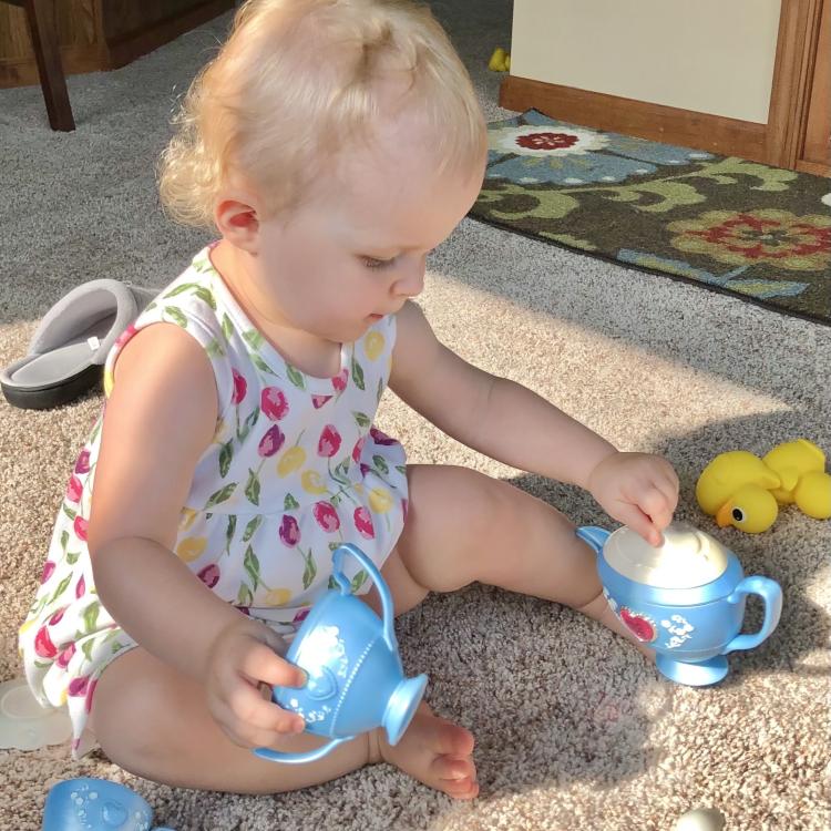 baby girl sits on floor, holding plastic teacup, in sunshine