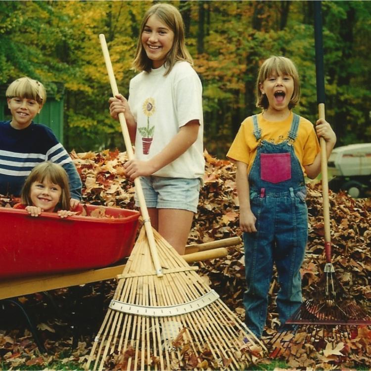 three kids rake leaves with one in wheelbarrow