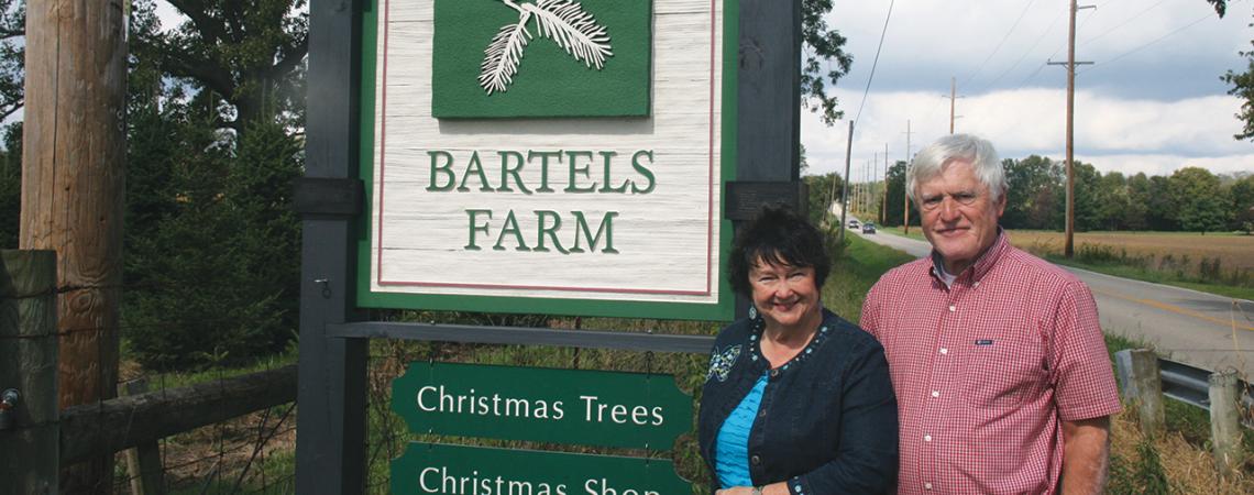 Judy and Steve Bartels smile next to the sign for their farm.