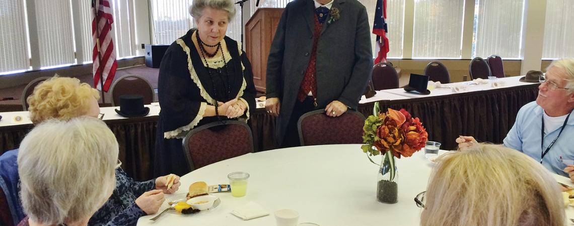 A group sits around a table as two impersonators visit with them.