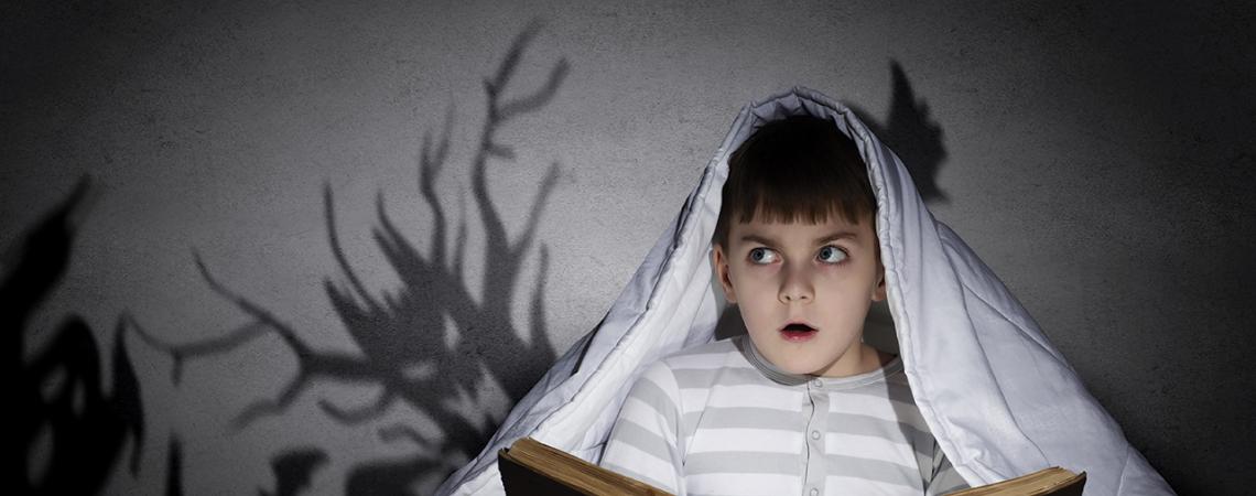 A boy reads under a blanket while scary shadows surround him.